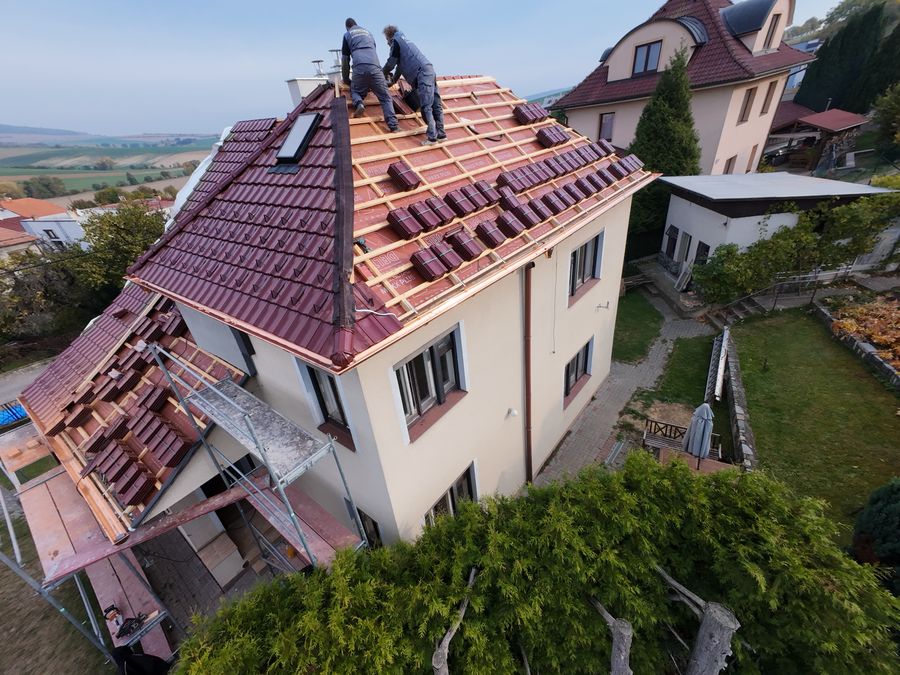 Pohled z dronu: dva pokrývači u komína na hřebeni, tašky rozložené na latích.